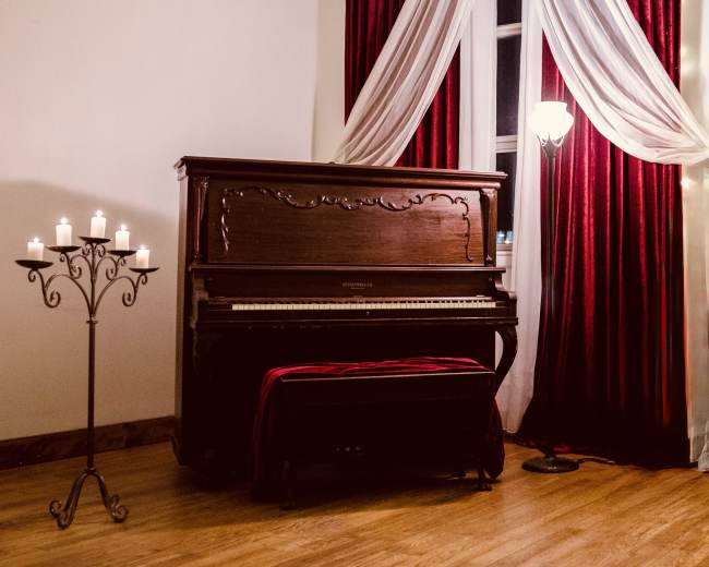 A vintage piano is situated against a wall with red curtains, accompanied by a candle holder and a floor lamp, creating a cozy atmosphere.