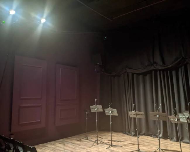 A dimly lit performance space with black curtains and several music stands arranged in front of them.