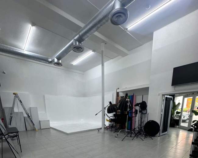 The image shows a spacious photo studio with a white backdrop, lighting equipment, and various props arranged around the room.