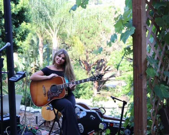 A woman sits outdoors on a chair, playing an acoustic guitar in a garden setting.