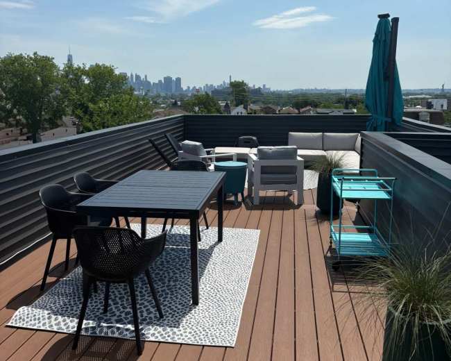 The rooftop terrace features a dining table, chairs, and lounge furniture, overlooking a city skyline under a clear blue sky.