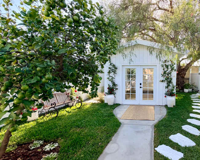 A white cottage with large glass doors sits at the end of a stone path lined with stepping stones, surrounded by green grass and trees.