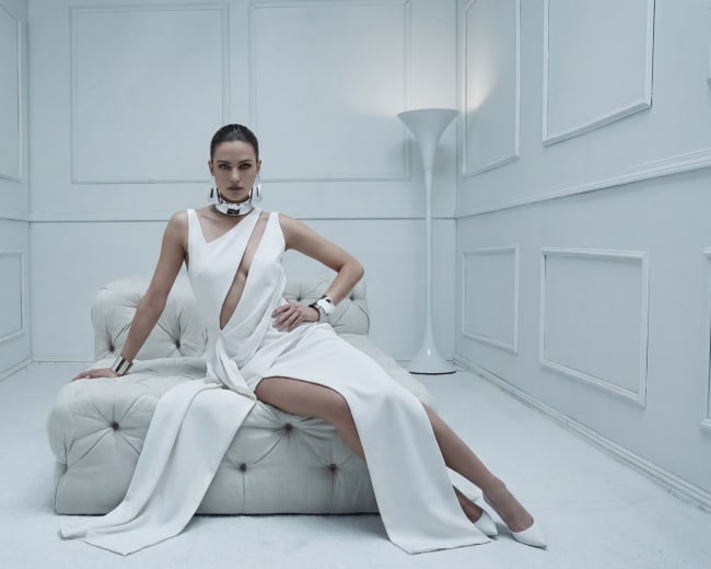 A woman in a white dress sits on a white couch in a minimalist, well-lit room.