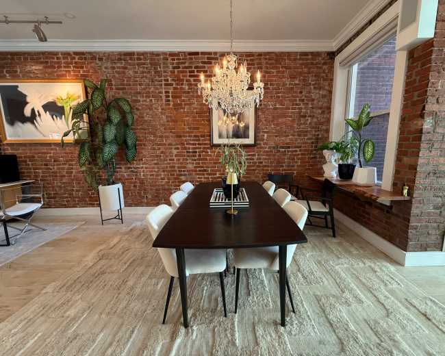 The image shows a dining area featuring a dark wooden table surrounded by white chairs, with a chandelier overhead and exposed brick walls.