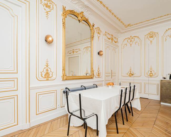 The image shows a richly decorated dining room with intricate gold accents on the white walls and a long table set for meals, accompanied by black chairs.