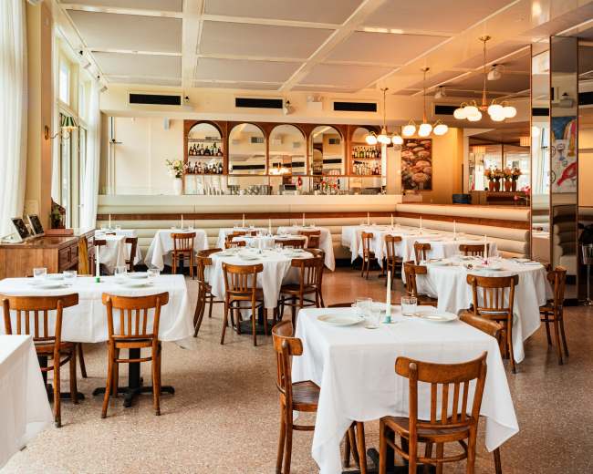 The image shows a spacious restaurant with wooden chairs and white tablecloths arranged around neatly set tables, illuminated by soft lighting and featuring a bar area in the background.