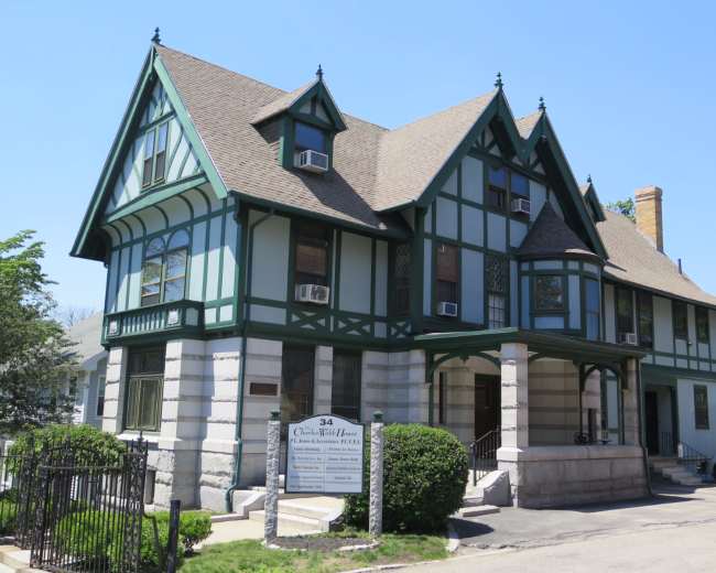 The image shows a large, three-story Tudor-style house with green wooden trim and a stone foundation, located near a street.