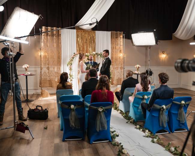A couple is exchanging vows during a wedding ceremony in a decorated venue, while guests sit in blue chairs.