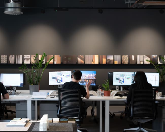 Three individuals work at desks with computers in a modern office space, surrounded by plants and a decorative wall of images.