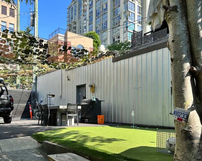 A small grassy area with a wooden bench and chairs in an urban setting, surrounded by buildings and a wall.