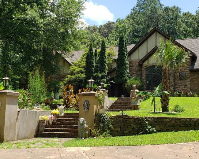 A large, stone-faced house is set atop a tiered lawn with palm trees and decorative statues lining the path to the entrance.