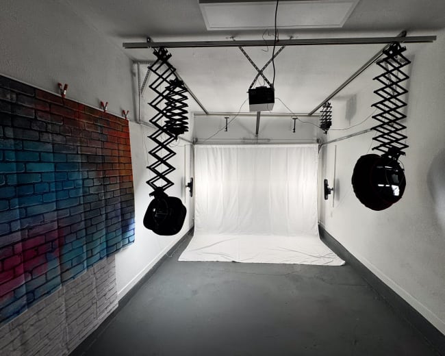 The image shows a photography studio with a white backdrop, two hanging lights, and a wall featuring a colorful mural of brickwork.