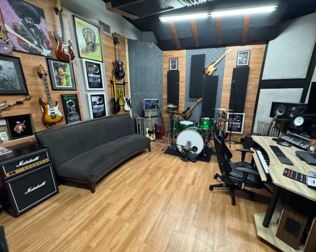 The image shows a music recording studio featuring guitars on the walls, a drum set, a keyboard, and a sofa, with soundproofing panels and artwork present.