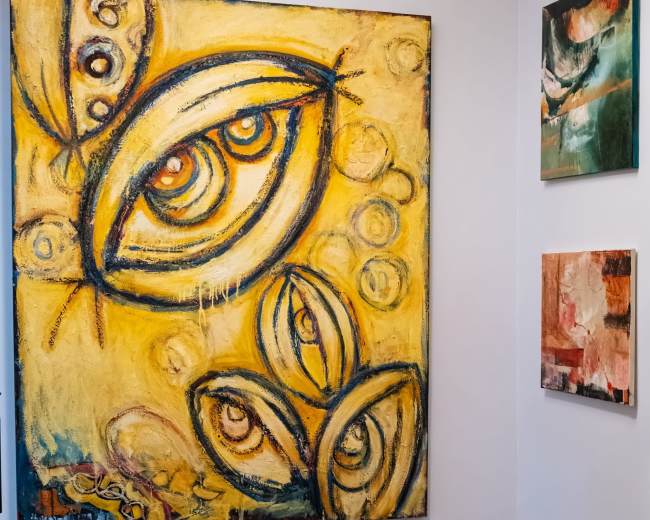 A large abstract painting featuring stylized eyes in yellow hues dominates the wall, accompanied by two smaller artworks on the adjacent wall beneath a starburst light fixture.