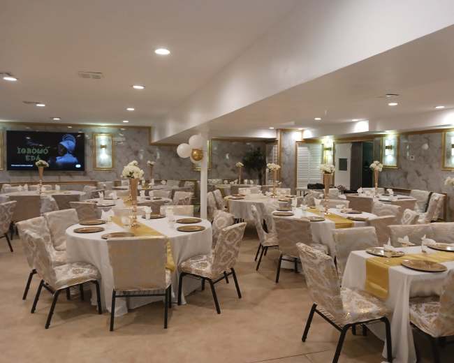 A banquet hall is arranged with neatly set tables featuring white tablecloths and gold accents, surrounded by elegant floral centerpieces.