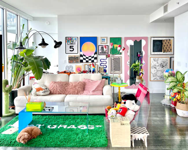 A brightly decorated living room features a white couch with pink cushions, a glass coffee table, colorful artwork on the walls, and various playful decor items.