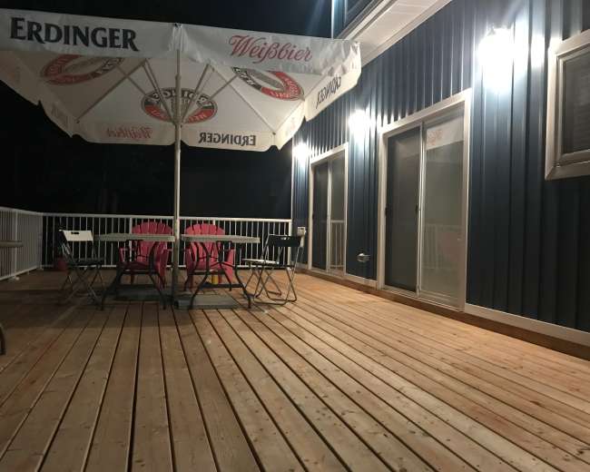 The image shows a wooden deck at night with a large umbrella and a table surrounded by chairs, illuminated by nearby lights.