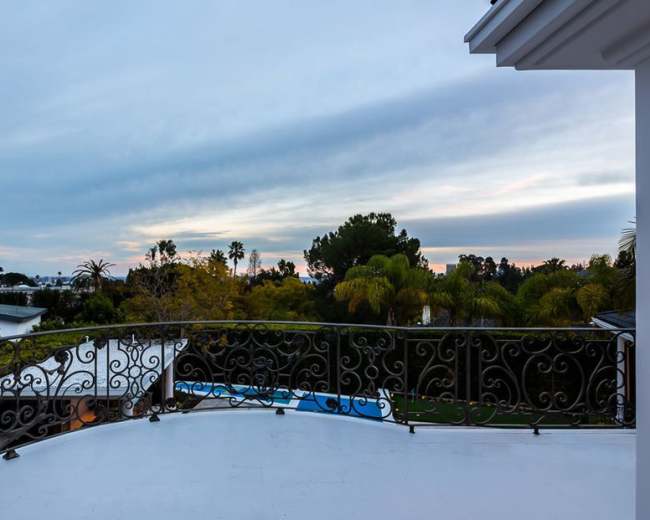 The image shows a balcony with an ornate railing overlooking a landscape of trees and a pool under a cloudy sky.