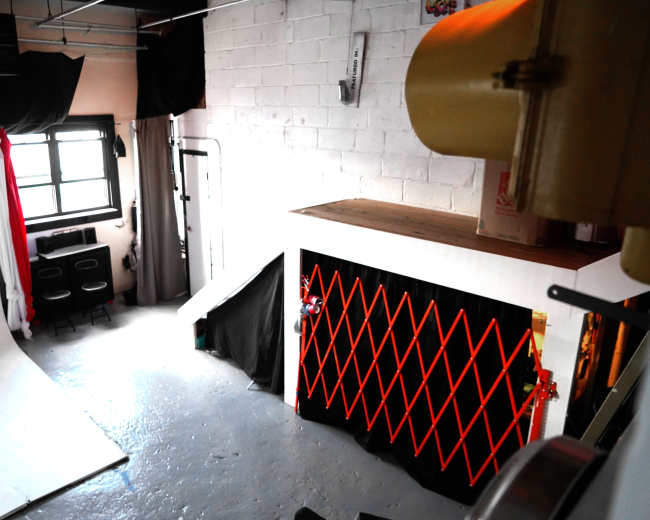 The image shows a photography studio with a white backdrop, a staircase, and an area enclosed by an orange safety gate.