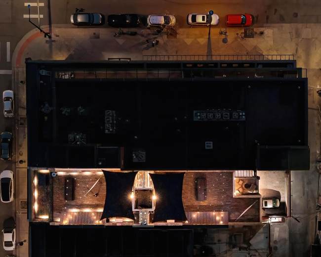 An aerial view shows a series of rooftops with parking spaces and vehicles, illuminated by surrounding lights at night.