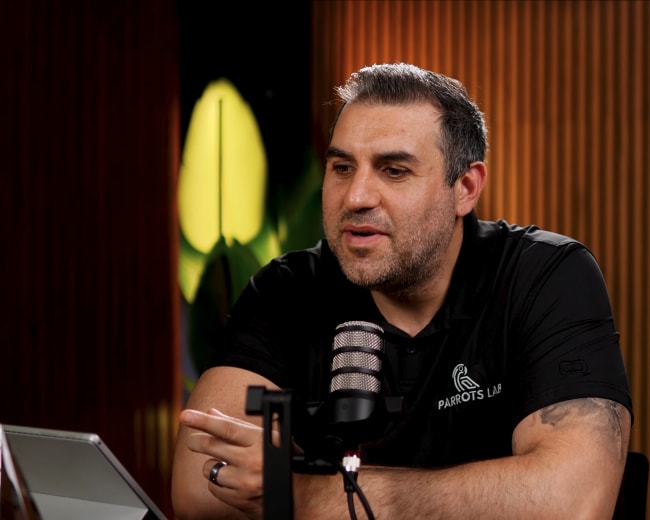 A man in a black shirt with a logo speaks into a microphone while seated at a table with a glass in front of him.