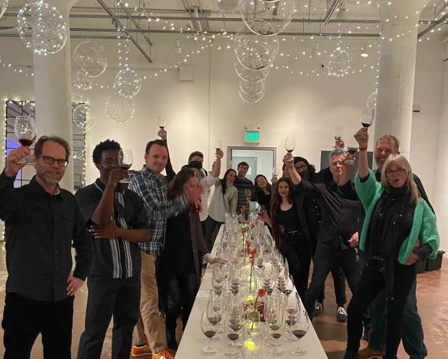 A group of people are toasting with wine glasses around a long table decorated with lights and glass ornaments in a brightly lit room.