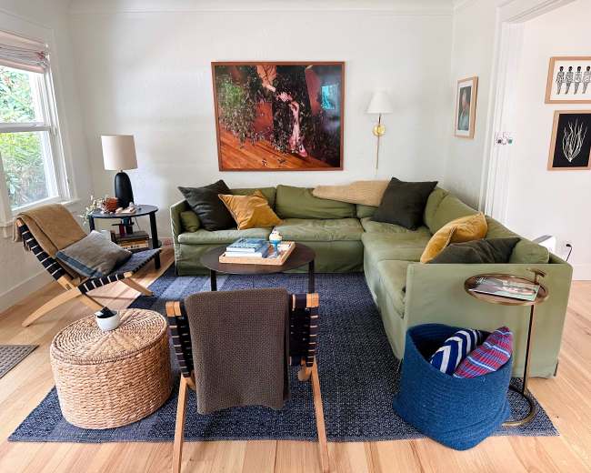 The image shows a cozy living room with a green sectional sofa, a black table in the center, a woven basket as a side table, and various decorative accents on the walls.