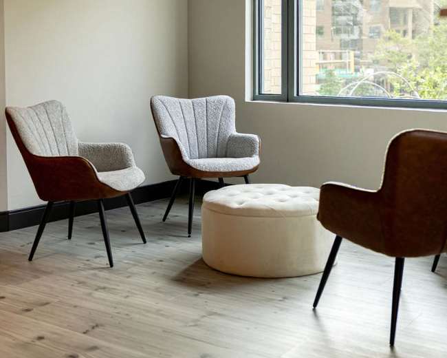 The image depicts a corner of a modern room with three upholstered chairs and a round ottoman, set near a large window overlooking a cityscape.
