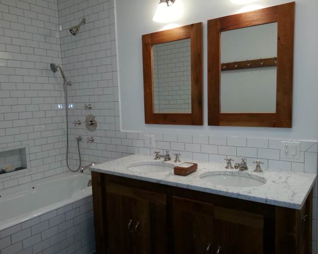 The image shows a bathroom featuring a white subway tile shower and bathtub, dual sinks with a marble countertop, and two wooden-framed mirrors hanging above.