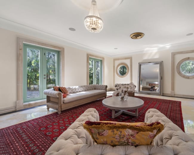 The image shows a spacious living room with light-colored walls, a circular mirror, a round coffee table, and two elegant sofas, all set on a patterned rug.