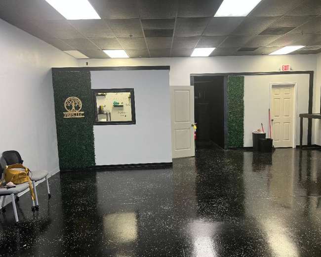 The image shows a spacious, mostly empty room with black floors, white walls, and a decorative green wall panel featuring a logo on one side.