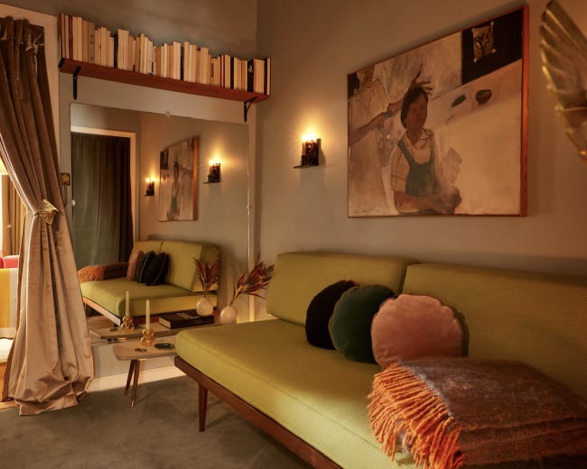 The image shows a cozy room with a green sofa, a painting on the wall, shelves of books, and warm lighting.