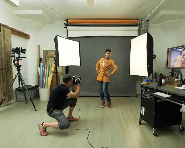 A photographer is capturing images of a model in a studio setting with softbox lights and a computer displaying the live feed.