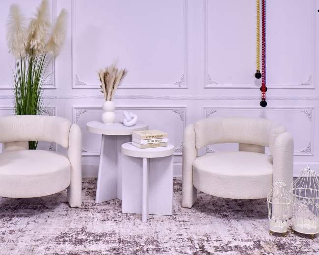 The image features two cream-colored chairs flanking a pair of modern side tables, positioned on a textured rug with decorative elements such as a vase and dried plants in the background.