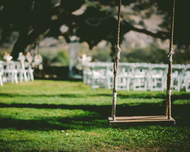 A wooden swing hangs from a tree, with a row of white chairs set up for an outdoor event in the background.