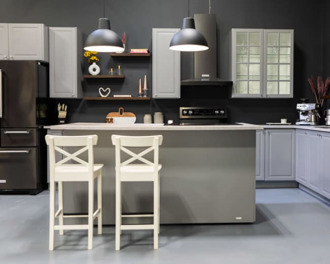The image shows a modern kitchen featuring gray cabinets, black appliances, and an island with two white stools.