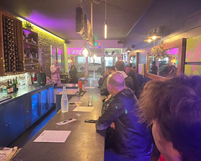 The image shows a dimly lit bar scene with patrons gathered at the counter and a bartender attending to drinks in a vibrant setting.