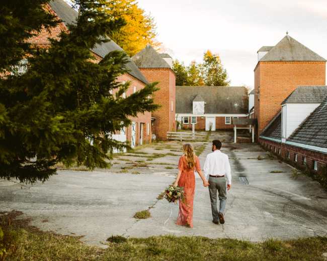 A couple walks hand in hand through a landscaped area with brick buildings in the background.