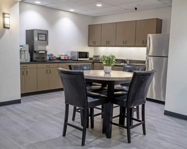 The image shows a modern kitchenette area with a round table and four black chairs, featuring cabinetry, a microwave, and a refrigerator.
