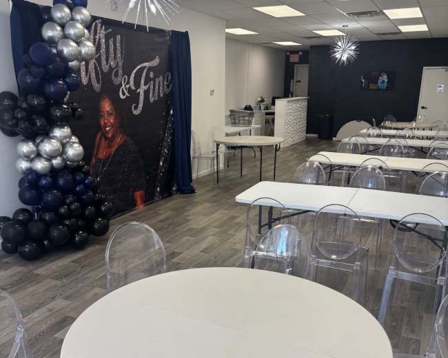 The image shows a decorated event space with several empty tables and a backdrop featuring a black and silver balloon arrangement next to a large portrait.