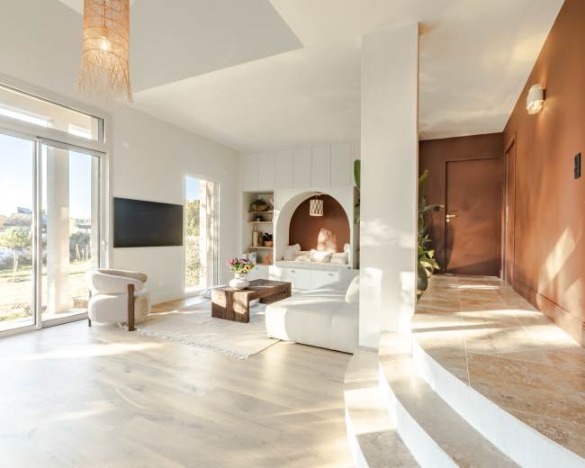 The image shows a modern living room with large windows, light wooden floors, and a circular seating area next to a wall with a geometric design.