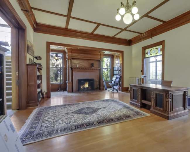 The image shows a spacious room with a wooden desk, a fireplace, and large windows, featuring a patterned area rug on the wooden floor.