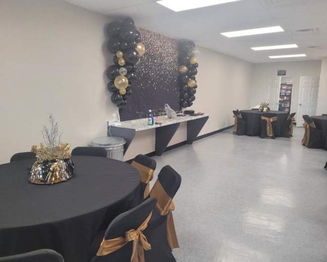 The image shows a decorated event space with black tables, gold accents, and a wall adorned with black and gold balloons.