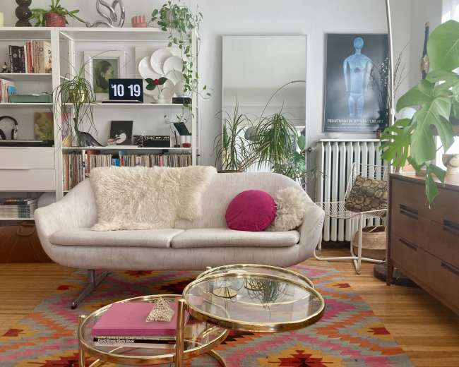 The image shows a contemporary living room with a light-colored couch, a round coffee table, various houseplants, and a bookshelf filled with decorative items and books.