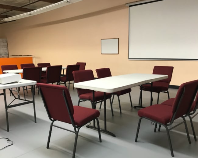 A meeting room is arranged with several chairs and tables, featuring a projector and a blank screen.