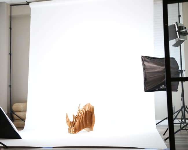 The image shows a wooden sculpture of a rhinoceros positioned against a bright white backdrop in a well-lit studio setting.
