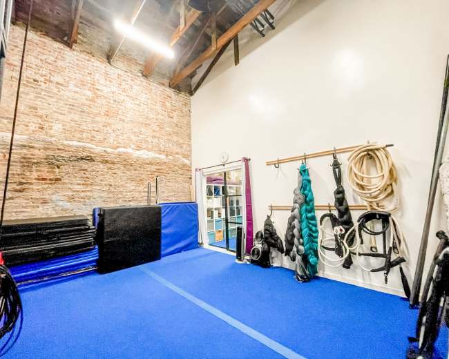 A training space featuring blue flooring, gym mats against a brick wall, and various equipment hanging on a rack.