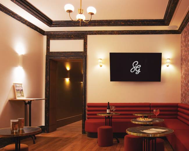 The image shows a stylish lounge area featuring red seating, a wall with floral wallpaper, and a television displaying a logo.