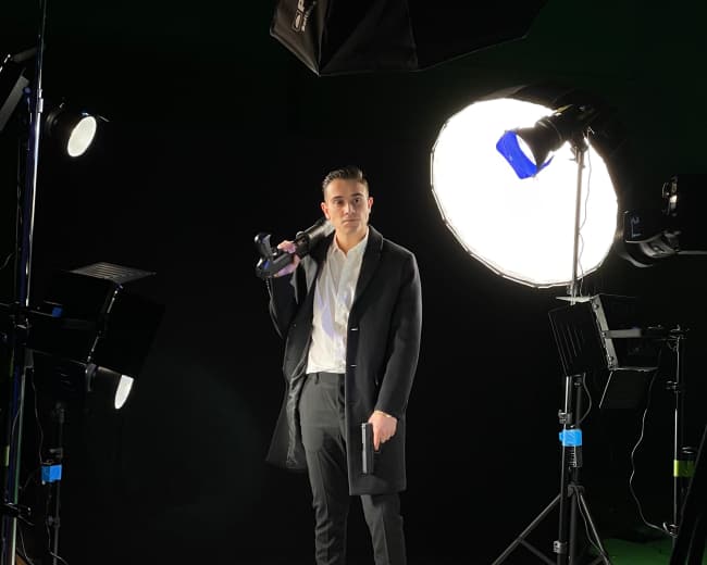 A man stands on a white backdrop holding a camera, surrounded by studio lighting equipment.