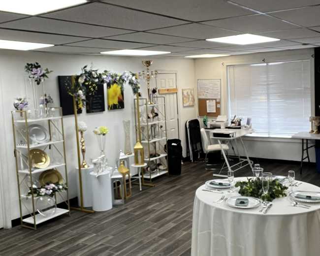 The image shows a decorated event space with a table set for a gathering, shelves displaying tableware, and various floral arrangements.